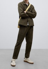 Person standing with arms crossed wearing olive green jacket and pants, white sneakers with gum soles, and a beige crossbody bag.