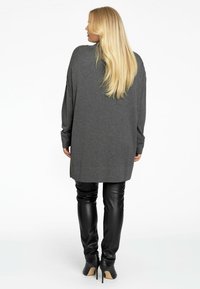 Grauer übergroßer Pullover mit langen Ärmeln, getragen über schwarzen enganliegenden Lederhosen und High Heels, zeigt eine glatte Textur und entspannte Form.