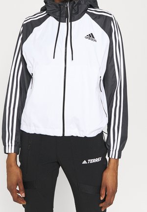 Persoon draagt een wit-zwart Adidas jack met capuchon met drie strepen op de mouwen en zwarte Adidas Terrex broek.