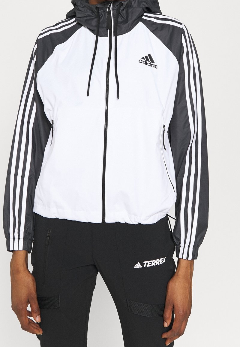 Personne portant une veste à capuche blanche et noire Adidas avec trois bandes sur les manches et un pantalon noir Adidas Terrex.