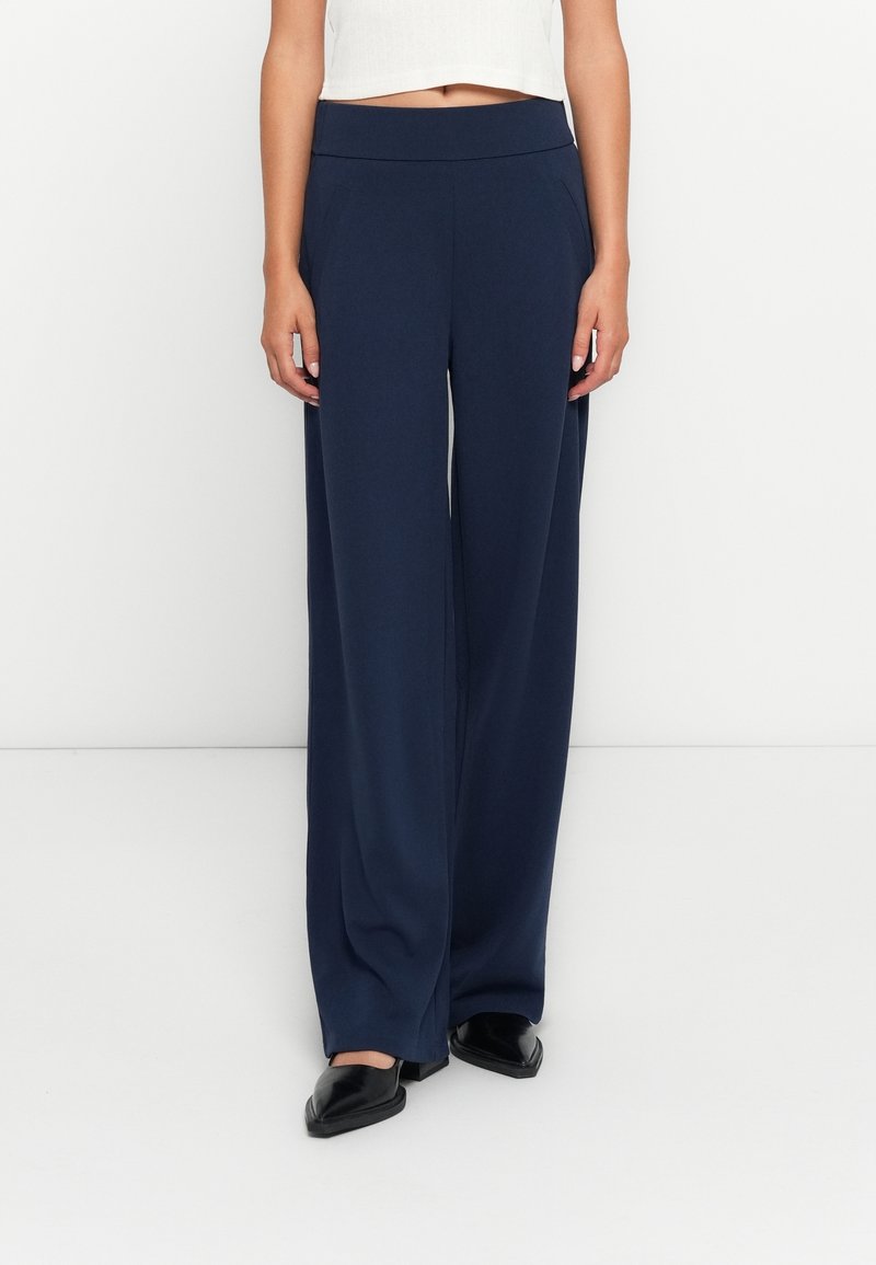 Femme portant un pantalon large taille haute bleu marine et des chaussures noires pointues, debout devant un fond blanc.