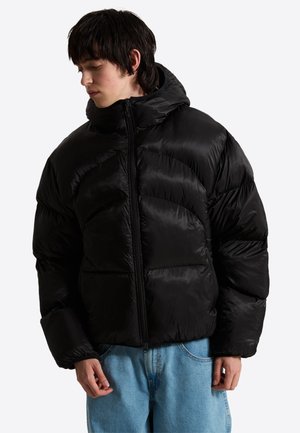 Giovane con giacca imbottita nera a collo alto e jeans blu, guarda verso il basso di lato su uno sfondo semplice.