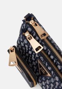 Taška s námořnickým vzorem a třemi zlatými zipy, s trojúhelníkovým logem značky na přední kapse na jednobarevném pozadí.