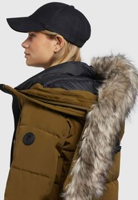 Tans puffjacka med svart dragkedja och avtagbar faux fur-huva. Har en svart cirkulär logotyp på ärmen och en svart keps.