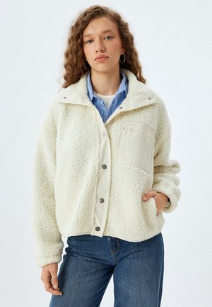 Veste en polaire crème, avec une fermeture à pression sur le devant, une poche poitrine et un tissu doux et texturé. Portée sur une chemise à rayures bleues.