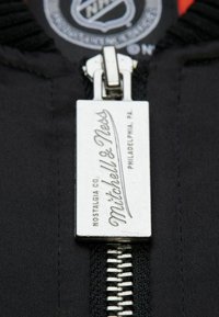 Metallreißverschluss mit einem rechteckigen silbernen Anhänger, auf dem "Mitchell & Ness" auf schwarzem Stoff eingraviert ist; mit strukturierter Oberfläche und Nähdetails.