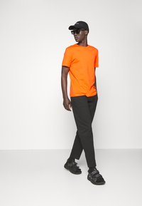 T-shirt di cotone arancione, cappello nero, occhiali da sole neri, pantaloni neri slim e sandali sportivi neri. Design semplice con accenti minimi.
