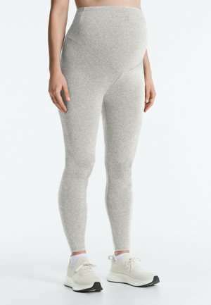 Personne enceinte portant des leggings de maternité gris clair et des chaussures de sport blanches, debout sur un fond blanc uni.