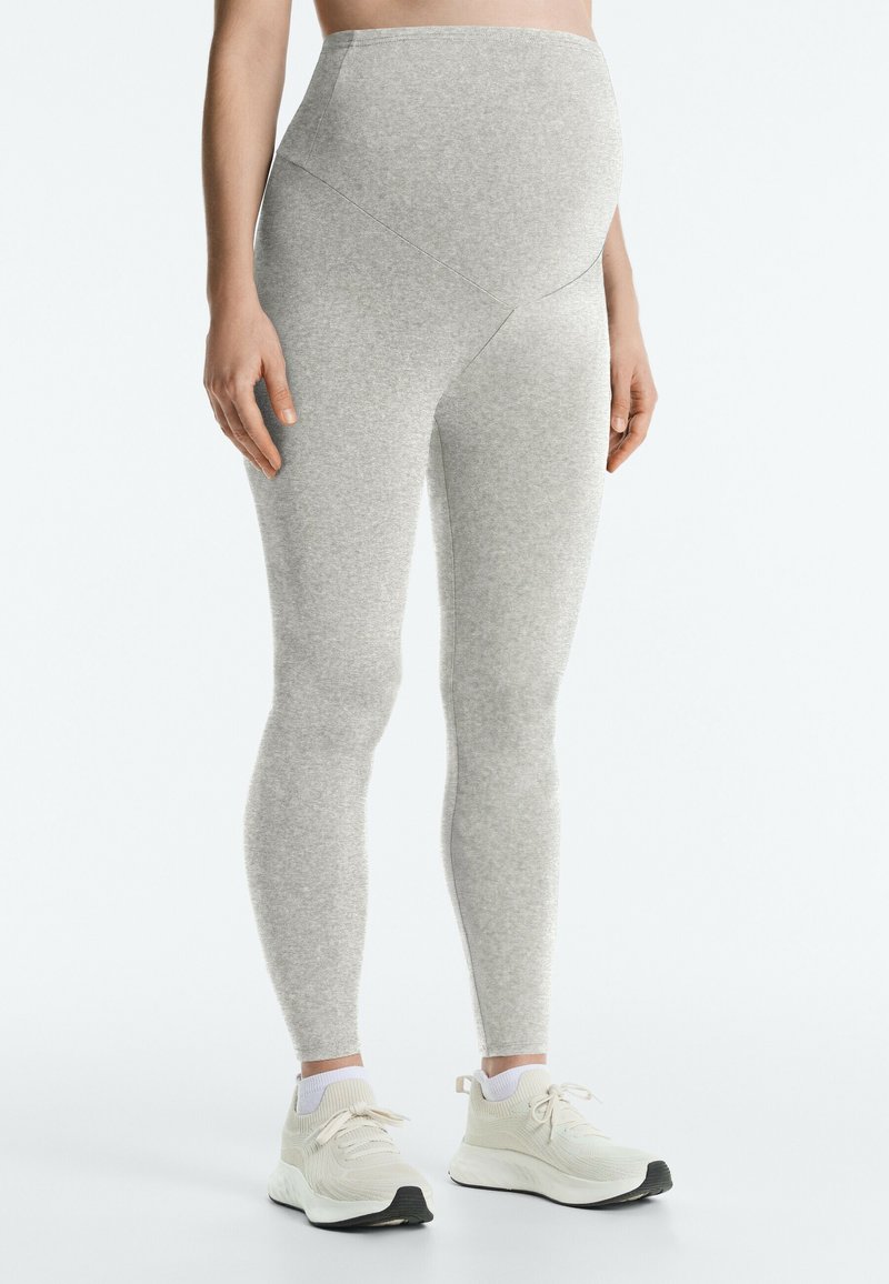 Personne enceinte portant des leggings de maternité gris clair et des chaussures de sport blanches, debout sur un fond blanc uni.