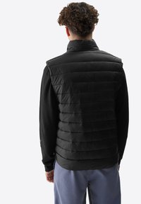 Schwarze ärmellose Steppweste mit horizontalem Steppmuster und hohem Kragen. Glatte, glänzende Oberfläche, kombiniert mit einem schwarzen Langarmshirt darunter.