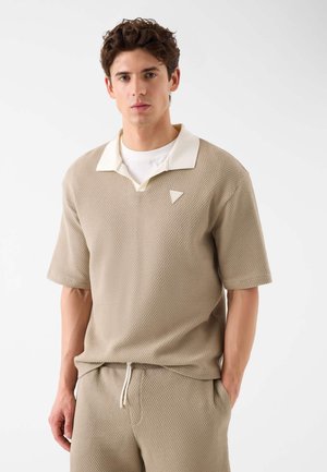 Junger Mann trägt beige texturiertes kurzärmeliges Hemd mit weißem Kragen und passende Hose mit Kordelzug, steht mit einer Hand in der Tasche.