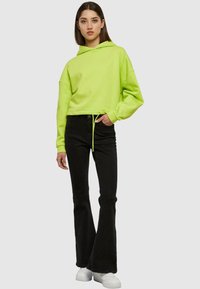 Felpa verde neon con cappuccio e lacci, vestibilità comoda e polsini a costine, abbinata a jeans neri a zampa d'elefante e sneaker bianche.