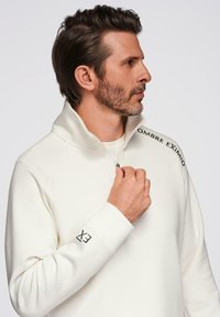 Sudadera de forro polar blanca con cuello alto, diseño de media cremallera y acento de texto negro que dice "OMBRE EXIMIO" en el hombro y "EX" en la manga.