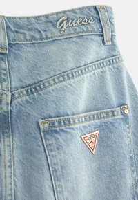 Jean en denim bleu clair avec le nom de la marque Guess brodé sur la ceinture et un patch logo triangulaire sur la poche arrière.