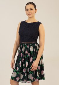 Granatowa sukienka z koronkową górą i spódnicą w kwiatowy wzór. Posiada dopasowaną talię i luźny krój w kształcie litery A. Teksturowany materiał z florystycznym wzorem.