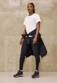 Valkoinen T-paita sinisellä logolla, mustat leggingsit sivupaneelilla ja mustat vaelluskengät. Musta takki sidottuna vyötärölle. Suora, moderni muotoilu.