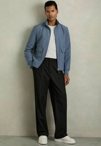 Modra zip-up jakna z dvema sprednjima žepoma, v kombinaciji s črnimi ohlapnimi hlačami in belimi športnimi copati. Gladek material in sproščen kroj.
