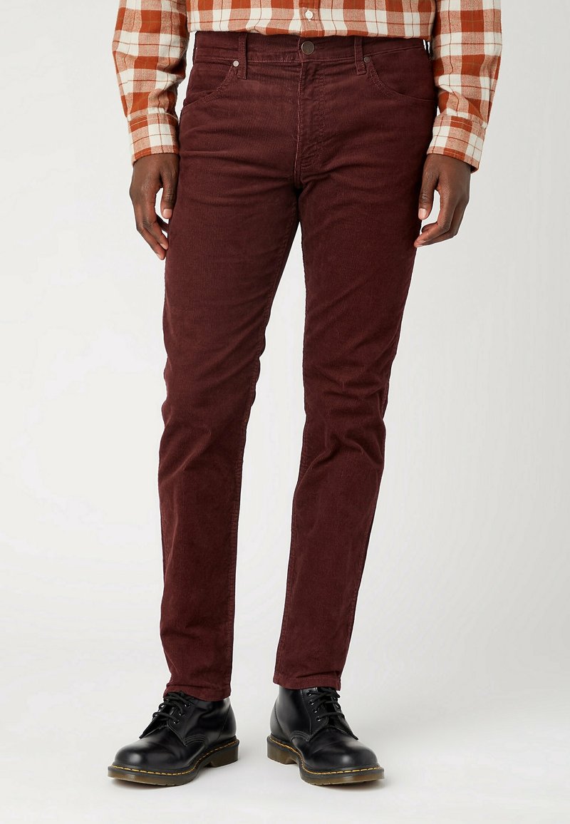 Homme portant un pantalon en velours côtelé rouge foncé, des chaussures en cuir noires à lacets et une chemise à manches longues à carreaux marron et blanc, debout sur un fond blanc.