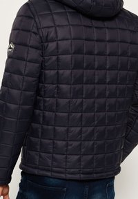 Tikkipintainen tummansininen toppatakki hupulla, jossa on teksturoitu ruudullinen kuvio ja vasemmassa olkapäässä logopläjäys.