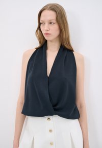 Blusa senza maniche blu navy con scollatura a V profonda e davanti drappeggiato, realizzata in tessuto morbido. Abbinata a una gonna bianca a vita alta con bottoni dorati.