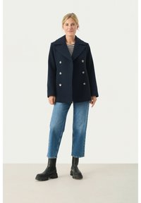 Marinblå ull peacoat med bred slitskrage och dubbelknäppning av silverknappar. Kombinerad med raka blå jeans och svarta stövlar.