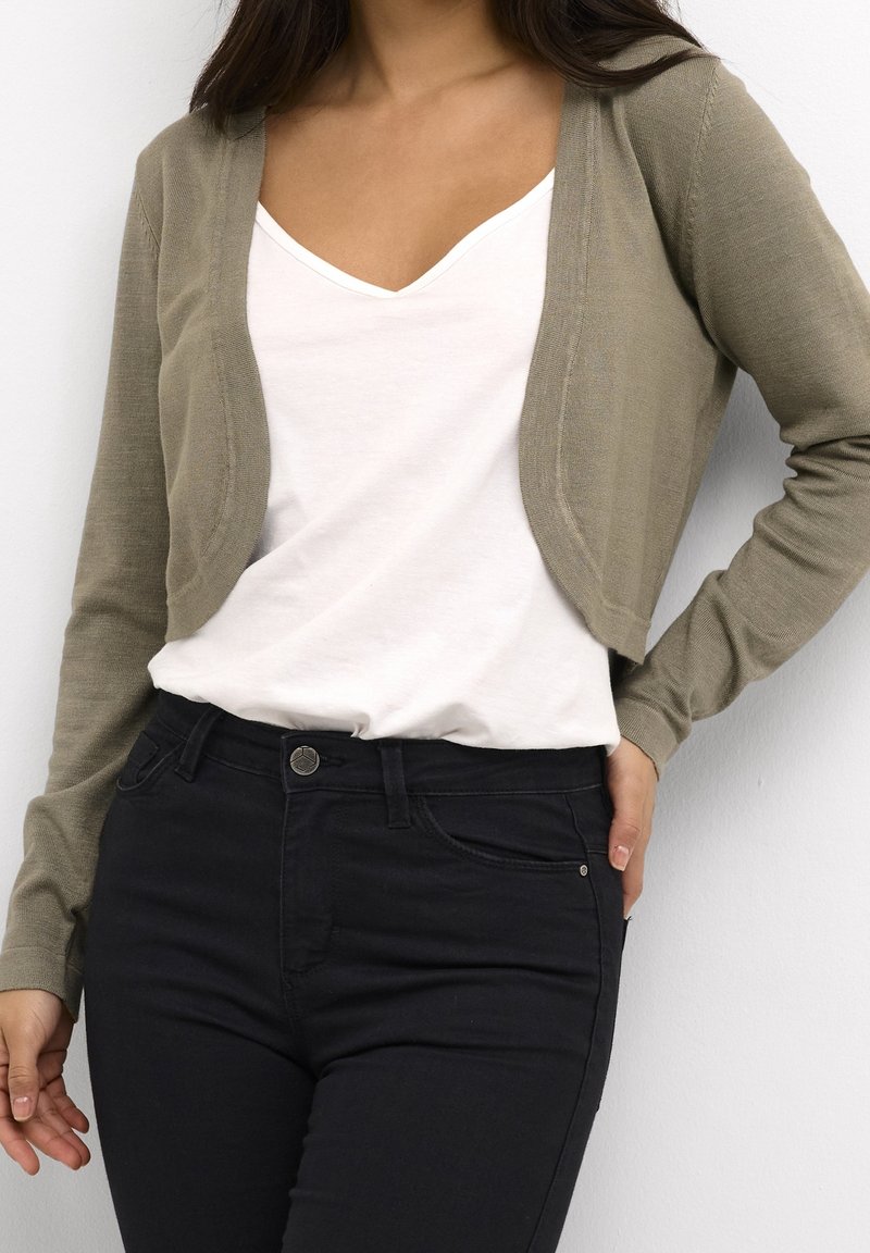 Cardigan court vert olive à manches longues porté sur un t-shirt blanc à col en V, associé à un jean noir ajusté. Texture lisse, design simple.