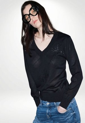 Vrouw met lang donker haar, grootzittende zwarte bril, een doorschijnende zwarte V-hals trui met subtiele patronen en een blauwe spijkerbroek met de handen in de zakken.