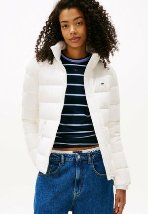 Witte pufferjas met een hoge kraag, ritssluiting en horizontaal gestikte uitstraling. Gecombineerd met een marineblauw gestreept shirt en blauwe jeans.