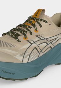 Μπεζ αθλητικό παπούτσι Asics με σκουροπράσινα κορδόνια με σχέδιο, πορτοκαλί οφθαλμούς, μπλε σόλα και μαύρο λογότυπο Asics στο πλάι.