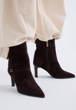 Botines de ante marrón oscuro con punteras puntiagudas, tacones delgados y cremalleras laterales, usados con pantalones beige.