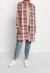 Karierter Mantel in Rosa-, Rot- und Türkistönen. Mit Kragen, Knopfleiste und langen Ärmeln. Trägt man über einem beigen Oberteil und Jeans.