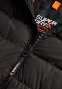 Primer plano de chaqueta acolchada negra con cremallera parcialmente abierta, mostrando el tirador de cremallera con la marca "Superdry" y la etiqueta interior con texto blanco y naranja.