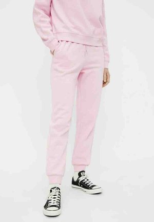 Person iført lyserøde joggebukser og matchende sweatshirt kombineret med sorte og hvide lavtop sneakers på hvid baggrund.