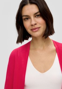 Rosa gerippter Cardigan mit offener Vorderseite, getragen über einem weißen V-Ausschnitt-Top. Das Design zeichnet sich durch weiche Texturen und eine lockere Passform aus.