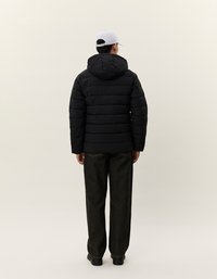 Sort polstret jakke med hætte, der har et quiltet design. Sammen med mørke jeans og en lys grå kasket.