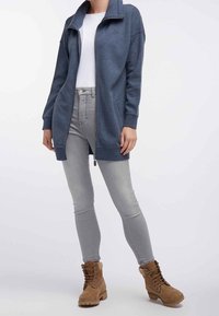 Giacca lunga blu con zip e colletto alto, indossata sopra una camicia bianca. Abbinata a jeans skinny grigi chiari e stivali marroni con lacci.