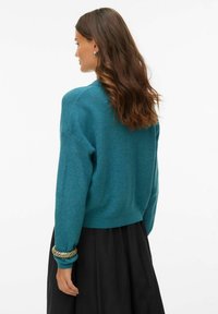 Turkusowy sweter na drutach o luźnym fasonie, z prążkowanym dekoltem i długimi rękawami. W zestawie czarna, kontrastująca spódnica i fakturowana bransoletka.