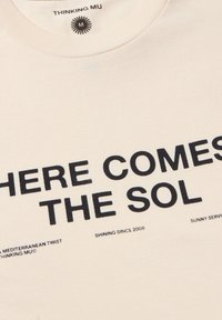 Camiseta de algodón color crema con texto negro en negrita: "AQUÍ VIENE EL SOL", con texto adicional debajo. Detalle de cuello redondo con acanalado.