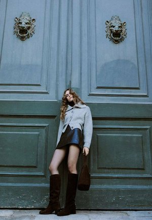Grauer Strickcardigan über einem schwarzen Leder-Minirock, kombiniert mit kniehohen schwarzen Stiefeln und einer kleinen braunen Handtasche, vor einer grünen Tür mit Löwenkopf-Türklopfer.
