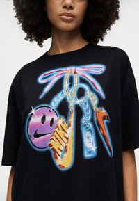 Γυναίκα που φοράει μαύρο T-shirt με πολύχρωμο σχέδιο από μπρελόκ σε σχήμα ροζ φιόγκου, χαμογελαστού προσώπου, λογότυπου Nike και Air tag.