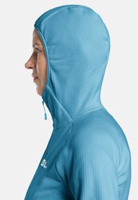 Veste à capuche bleu clair avec un motif texturé, dotée d'une fermeture éclair et d'une coupe ajustée. Montre le profil latéral de la silhouette à capuche.