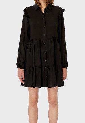 Vrouw gekleed in een zwarte, lange mouwen, gelaagde jurk met knopen aan de voorkant en ruches op de schouders tegen een effen achtergrond.