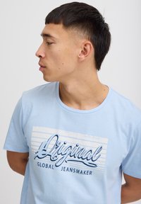 Lyseblå bomull T-skjorte med korte ermer, med et grafisk trykk som leser "Original Global Jeansmaker" i mørkeblå bokstaver.