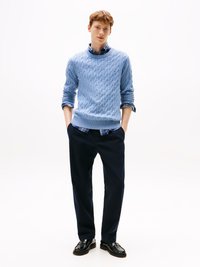 Suéter de malha em azul claro sobre uma camisa xadrez, combinado com calças escuras e sapatos de couro pretos. Design simples e limpo.