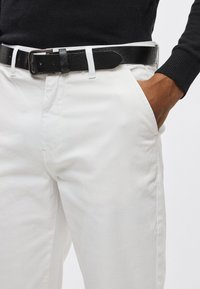 Białe bawełniane spodnie o prostym kroju, wyposażone w czarny skórzany pasek i boczne kieszenie. Pasek posiada metalową klamrę.