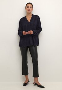 Blouse à manches longues bleu marine avec des rayures verticales argentées, col en v, détails volantés au cou, associée à un pantalon en cuir noir coupé et des escarpins à brides arrière.