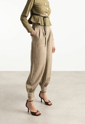 Vrouw draagt beige geplooide enkellange broek, olijfgroene top met knopen en zwarte riem, en bruine sandalen met hoge hakken, staand op een neutrale vloer.