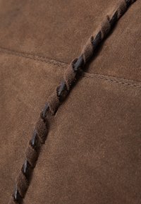 Bruin suede materiaal met een gestructureerd oppervlak, met decoratieve donkerbruine stiksels die langs de naad lopen en vakmanschapdetails benadrukken.