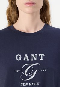 Marineblå T-shirt med rund halsudskæring og korte ærmer. Har hvidt trykt tekst: "GANT EST. 1949 NEW HAVEN" og et stiliseret "G."