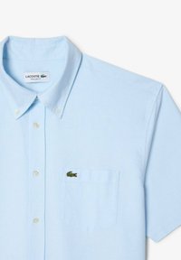Chemise col bleu clair, à manches courtes, design boutonné, présentant une poche poitrine avec un logo de crocodile vert brodé. Coupe régulière.